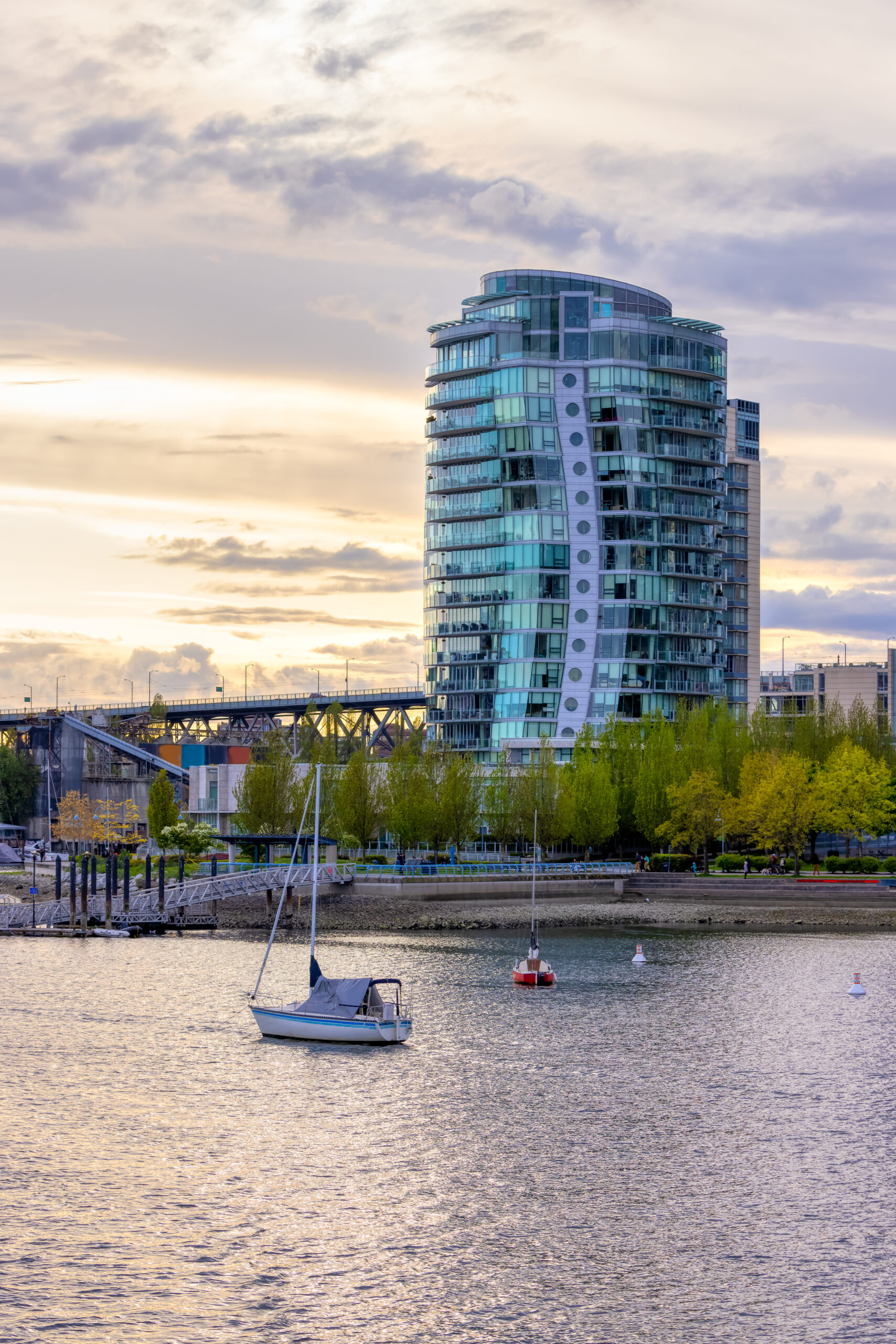 Modern glass high-rise condominium building overlooking Nanaimo harbour at sunset, with sailboats moored in foreground and Granville Street bridge visible behind, surrounded by spring trees