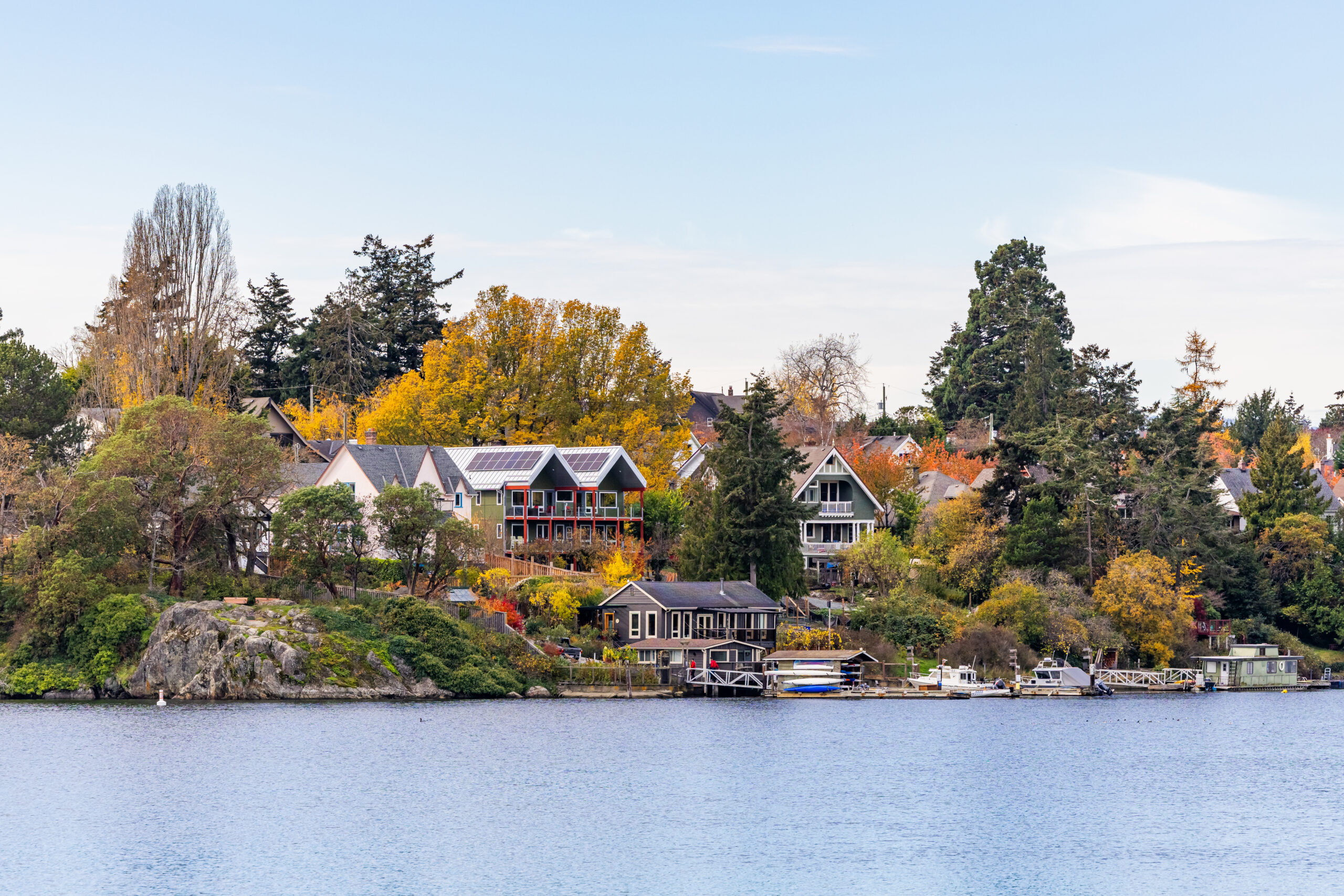 This image shows a scenic waterfront neighborhood during autumn. Several residential homes sit on a rocky hillside overlooking a lake or bay. The homes feature diverse architectural styles, including some modern designs with solar panels and traditional A-frame houses. Fall colors are prominent with yellow and orange foliage on the trees surrounding the properties